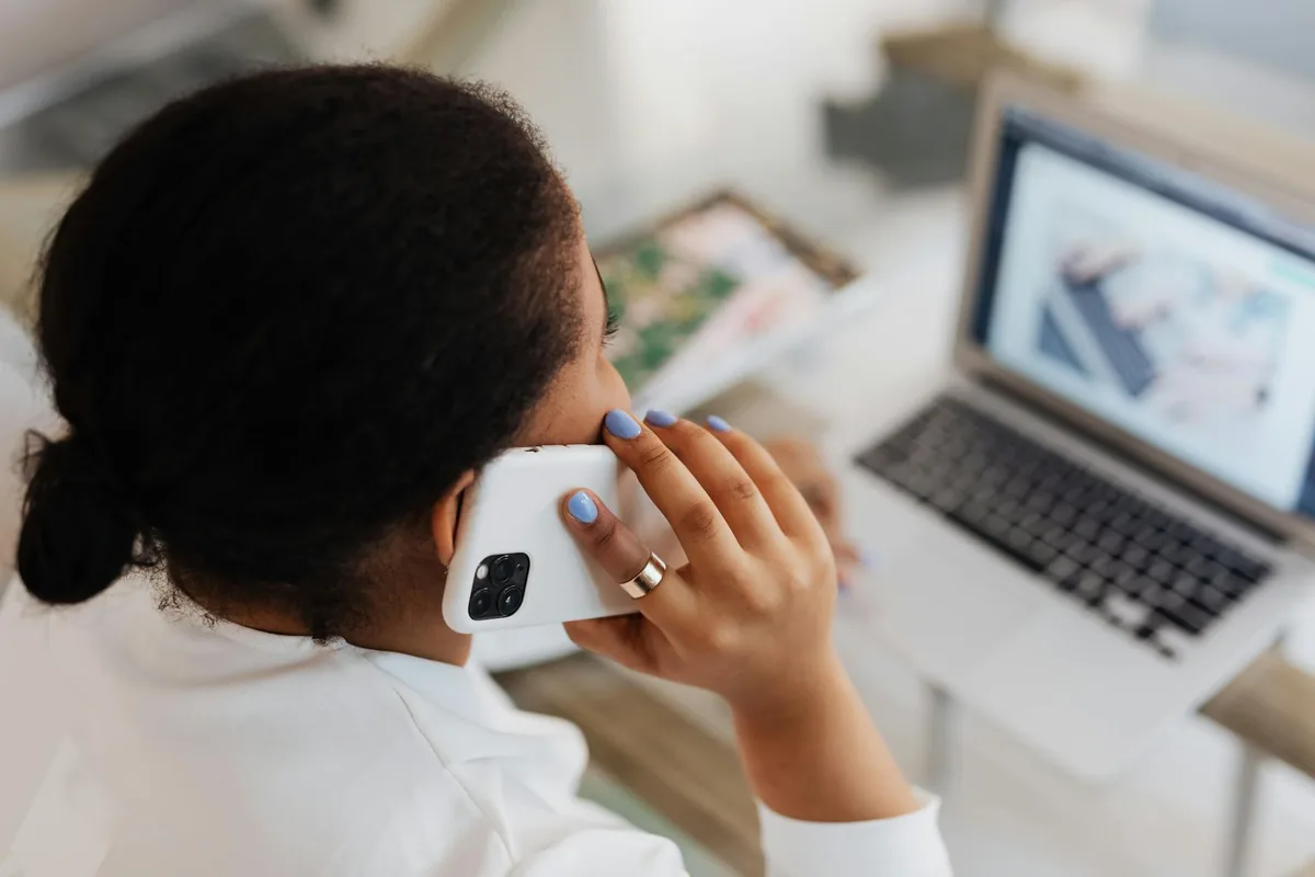 Mulher falando ao celular diante de um notebook aberto, em mesa de escritório