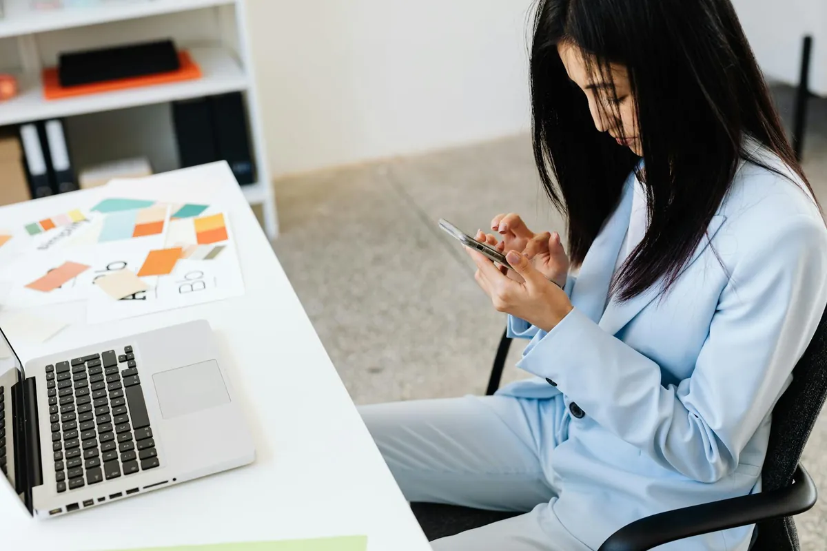 Profissional de blazer azul usando o celular ao lado de um laptop aberto numa mesa de escritório