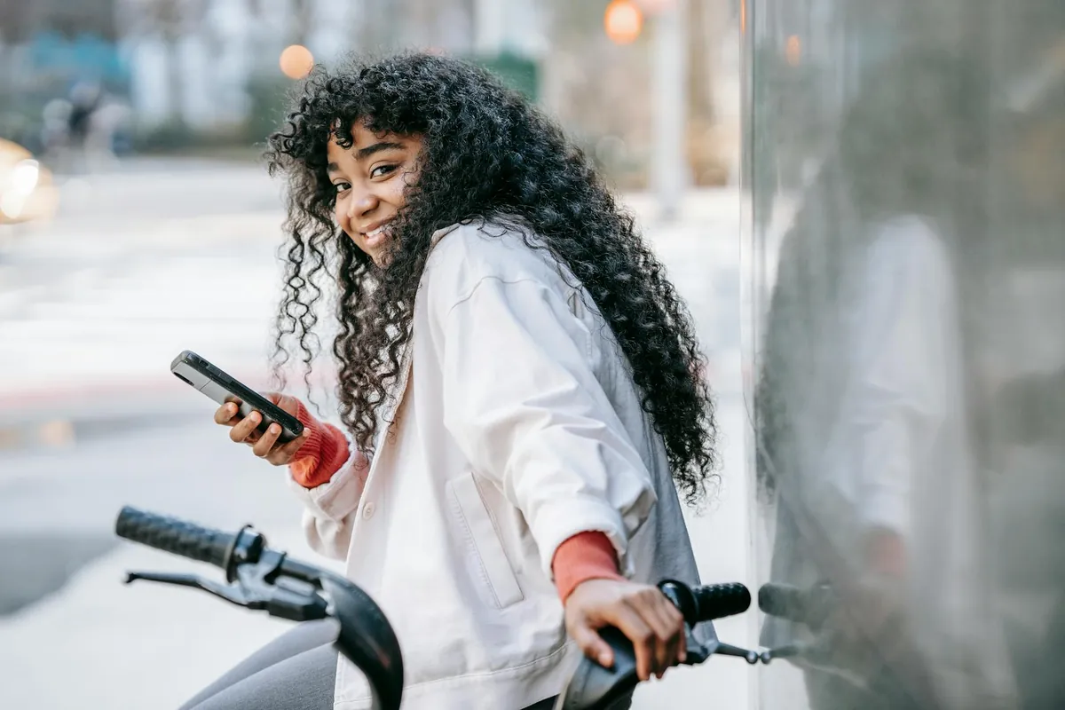 Jovem sorrindo na rua com o celular na mão, encostada numa bicicleta