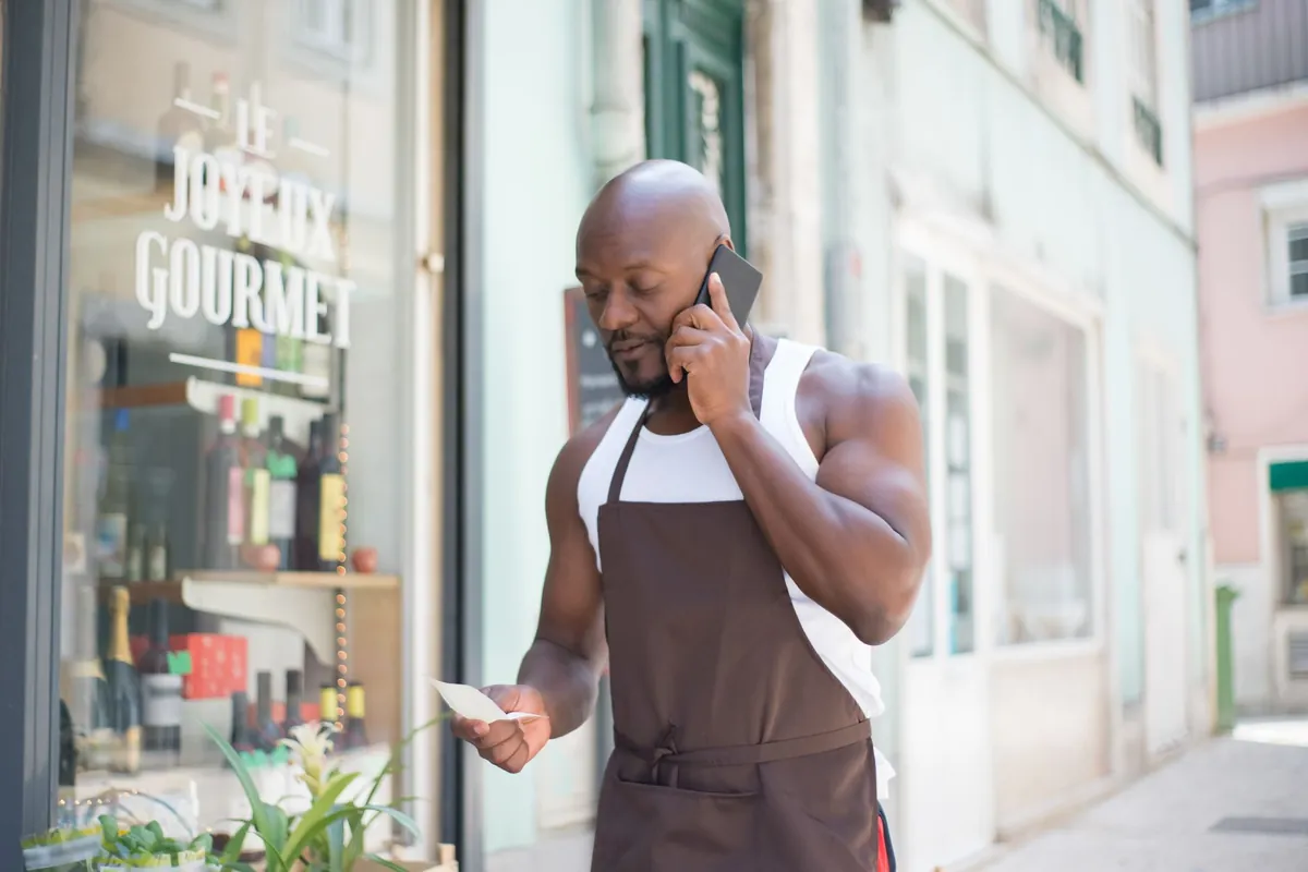 Comerciante de avental, na porta da loja, fala ao celular enquanto confere um papel na mão